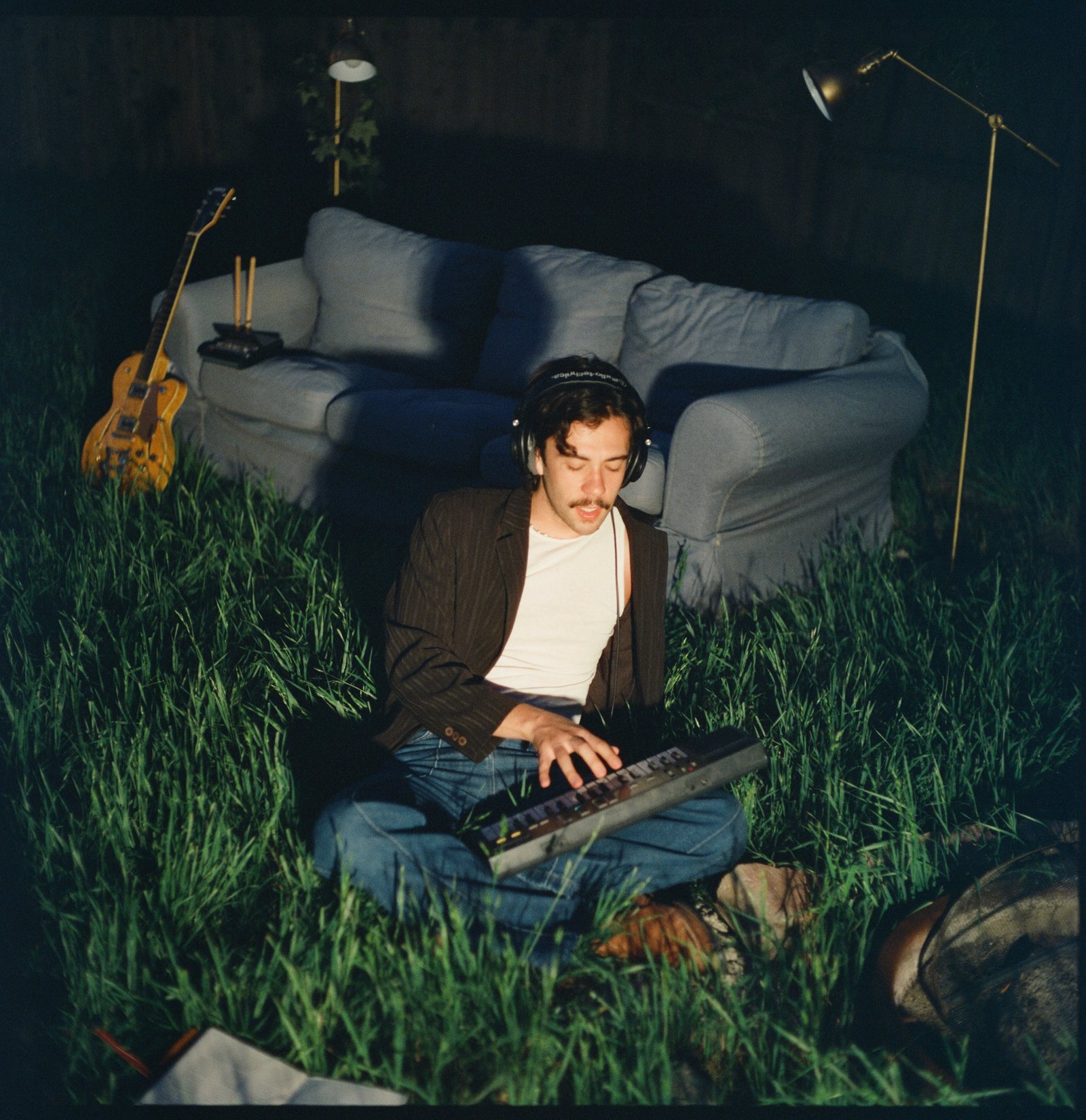 Seth Celdrán on a lawn at night in over-ear headphones, a keyboard across his knees, a couch, two floor lamps and a yellow Gretsch guitar behind him